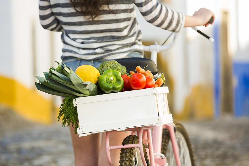 Kobieta prowadząca rower ze skrzynką na bagażniku wypełnioną świeżymi warzywami-  Jak kupować bardziej eko.