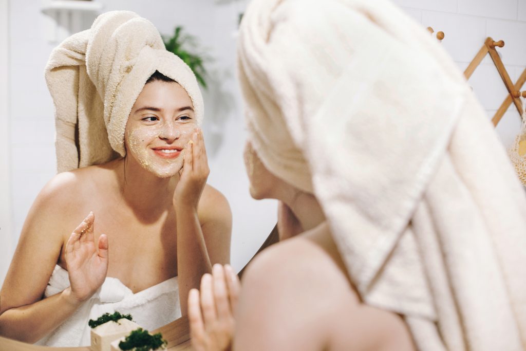 : Kobieta z ręcznikiem na głowie przeglądająca się w lustrze i nakładająca na twarz maseczkę – Domowe SPA